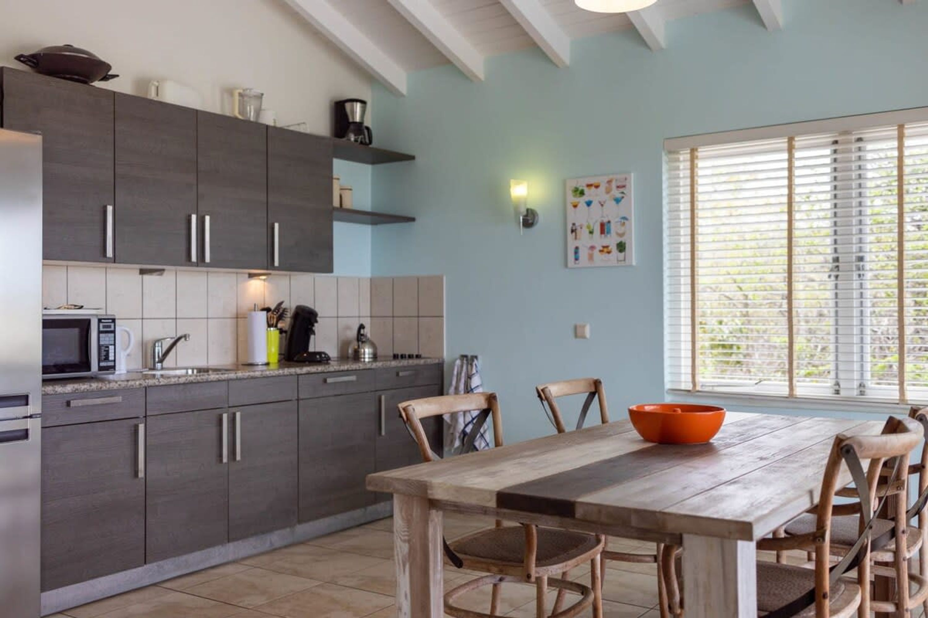 A modern kitchen with dark cabinets, a wooden dining table, and a large window with blinds.