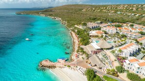 Une vue aérienne d’une station balnéaire avec une plage, des eaux turquoise et limpides, et une rangée de bâtiments blancs aux toits orange.