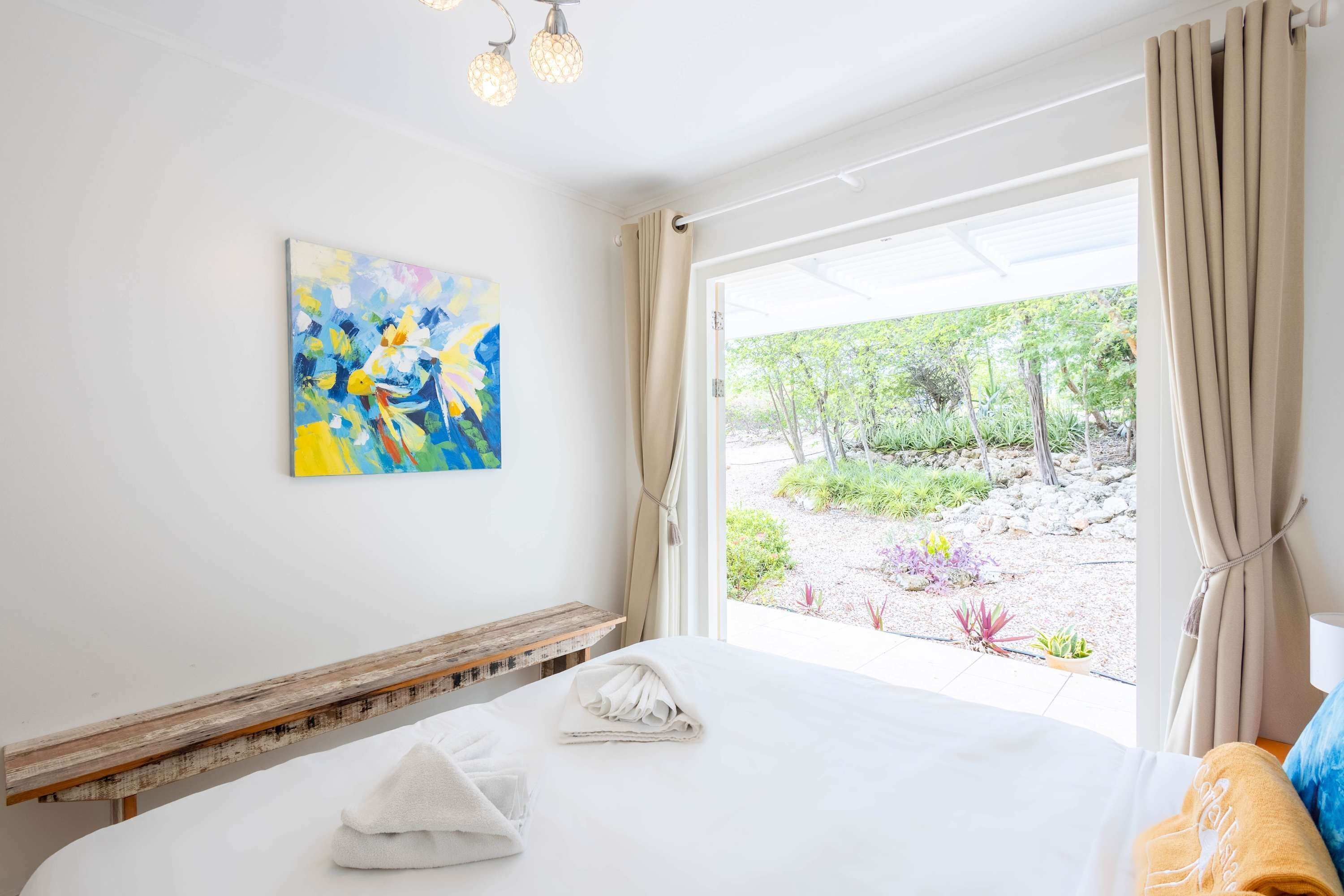 A hotel room with a bed, a painting on the wall, a window with curtains, and a view of an outdoor garden.