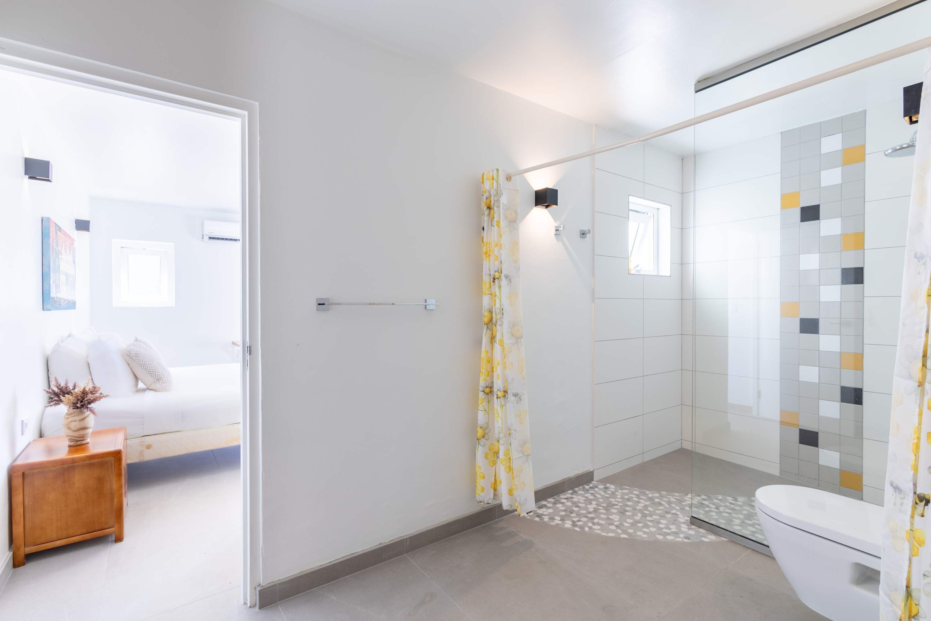 A modern bathroom with a glass shower enclosure, a toilet, and a small bedside table with a vase.