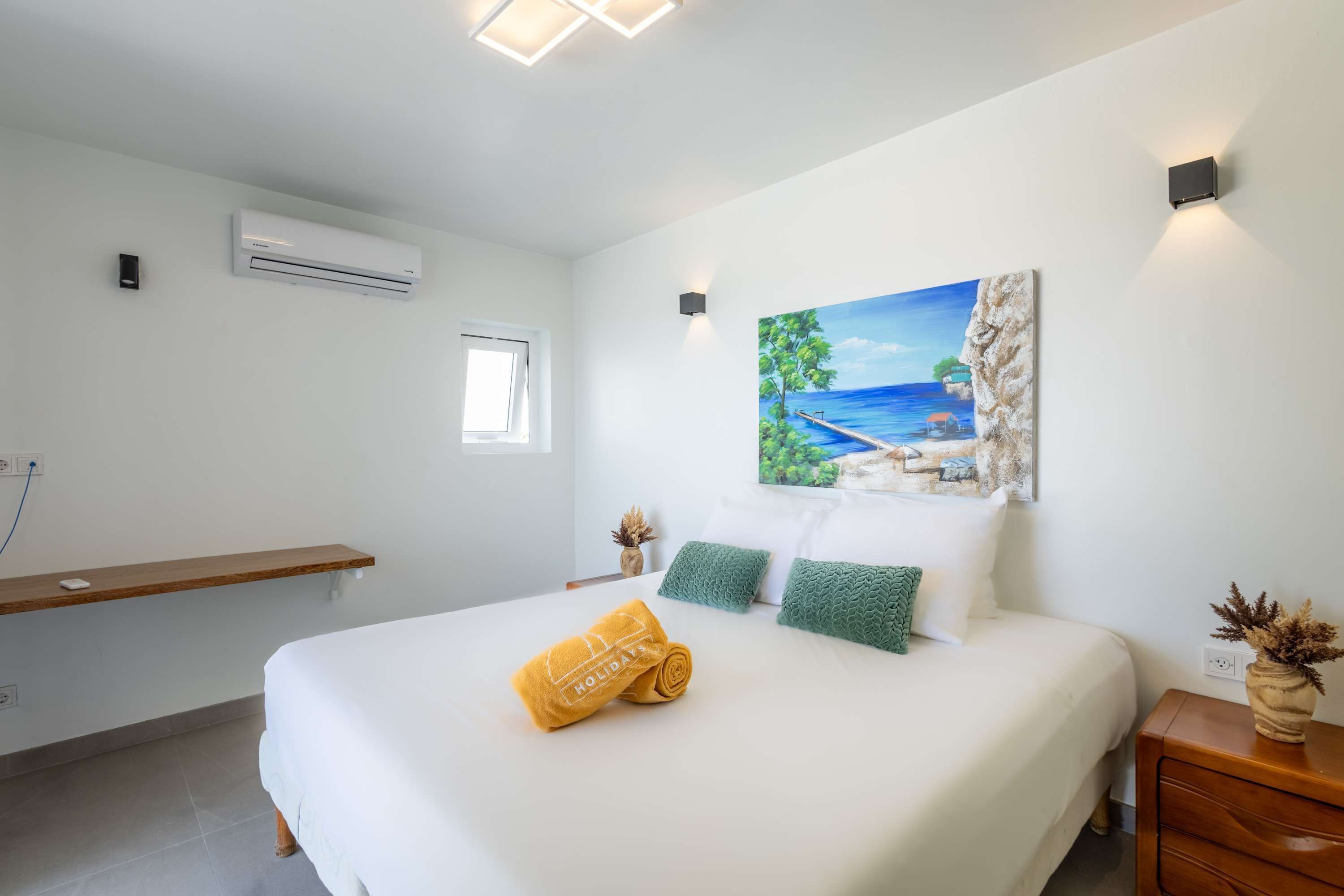 A hotel room with a bed, a painting of a beach scene, a window, and an air conditioning unit.