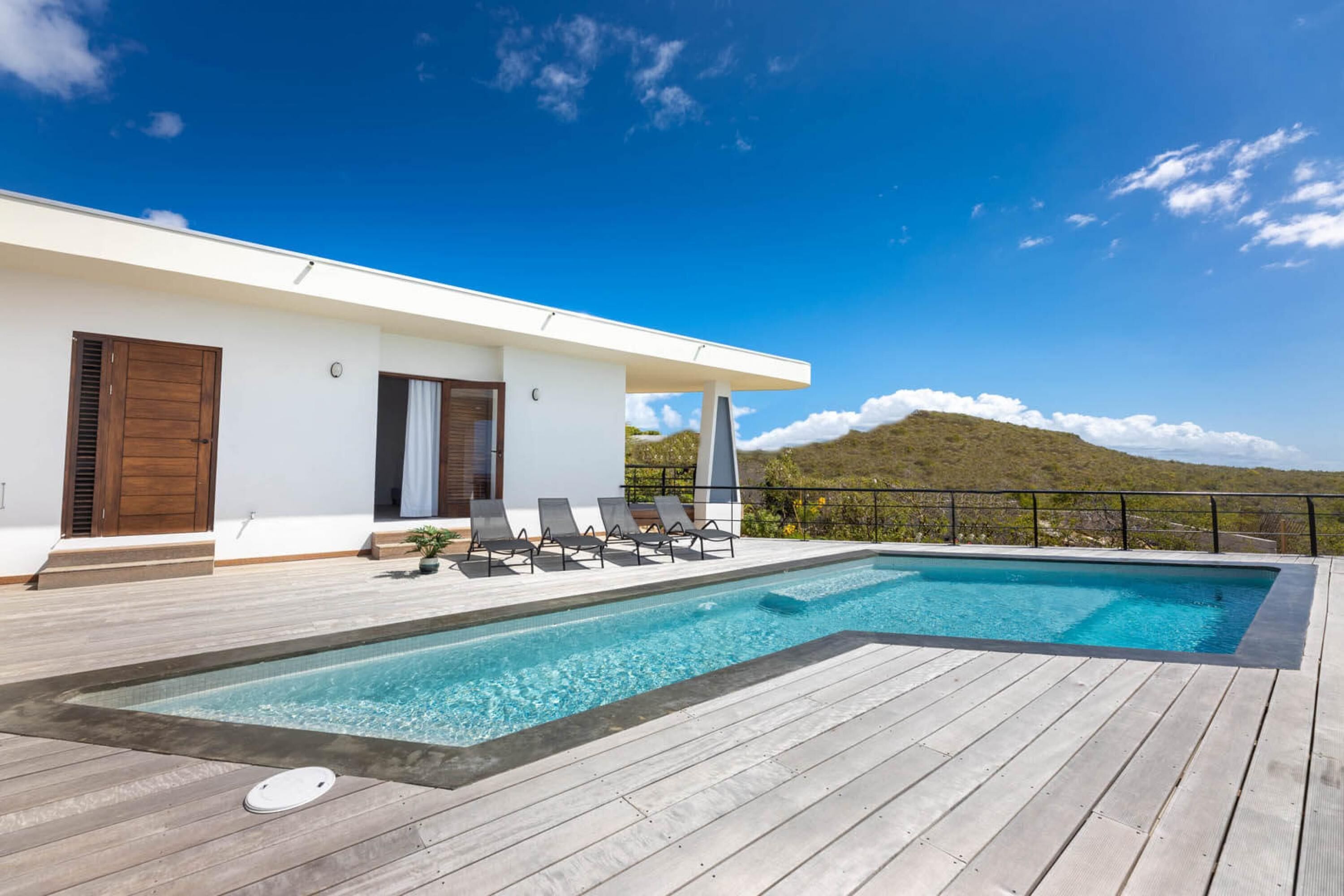 A modern pool area with a wooden deck, lounge chairs, and a view of a mountain.