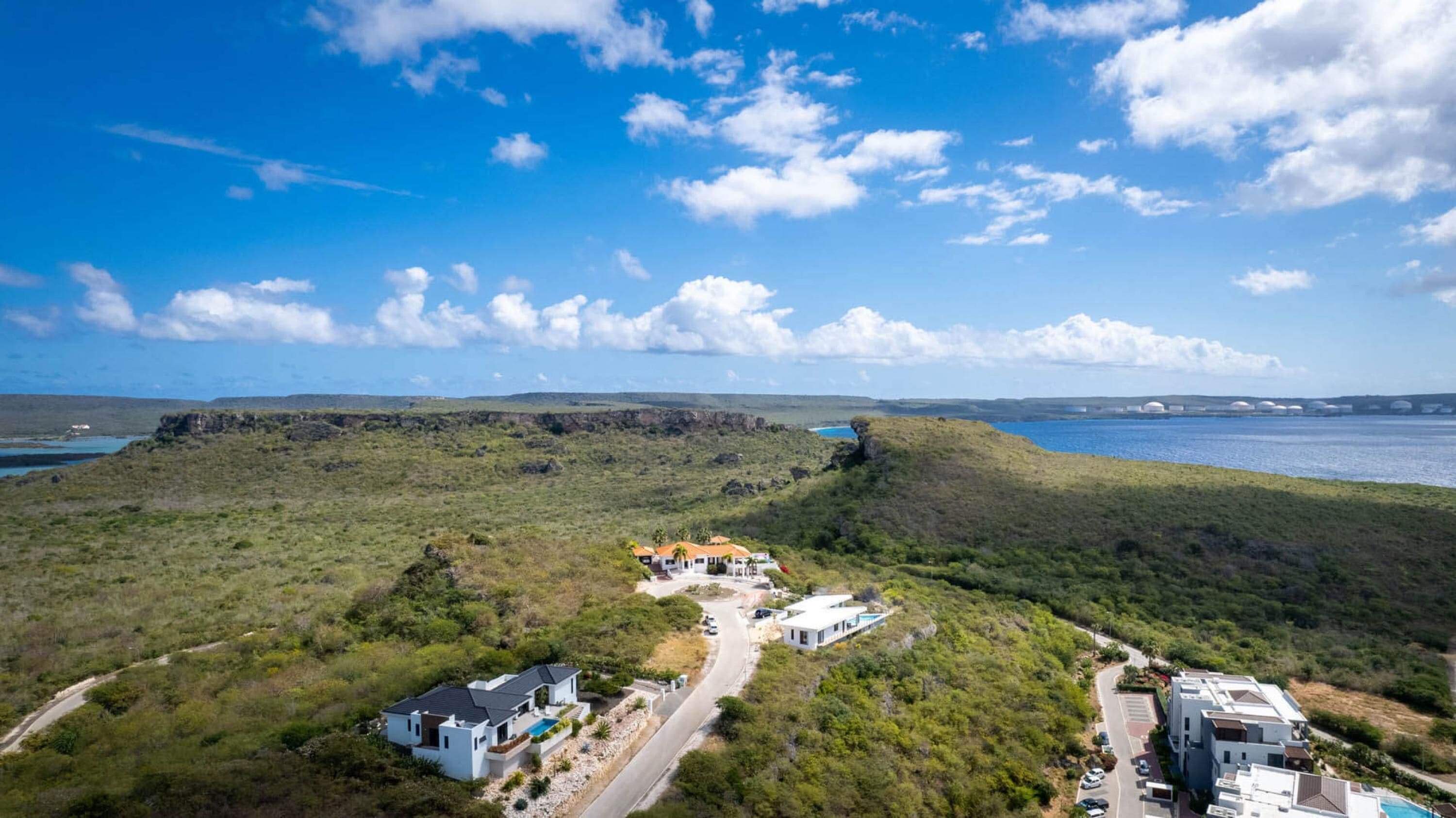 A coastal property with a swimming pool, a road, and a view of the sea and distant buildings.
