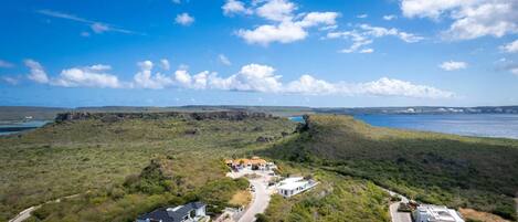 A coastal property with a swimming pool, a road, and a view of the sea and distant buildings.