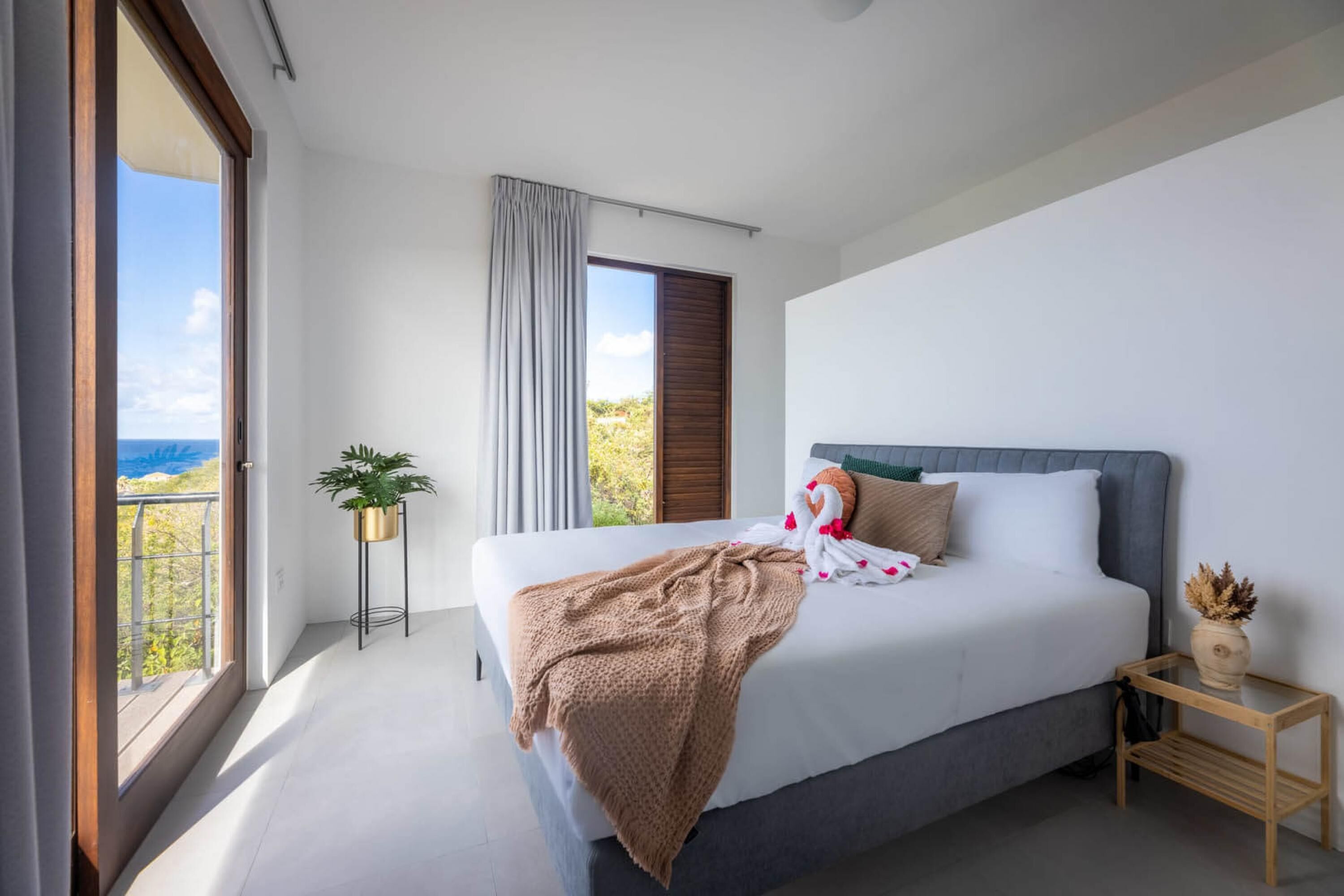 A modern bedroom with a large bed, a view of the outdoors, and a small side table with a plant.