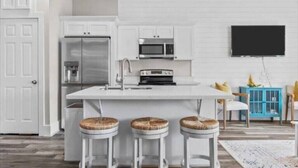 Een moderne keuken met witte kasten, een roestvrijstalen koelkast, een magnetron en een ingebouwde oven. Er staan drie barkrukken met gevlochten zittingen bij het kookeiland. Een flatscreen televisie is aan de muur boven de keuken bevestigd.