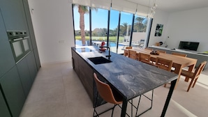 A modern kitchen with a large island, a dining area with wooden furniture, and a view of a swimming pool and palm trees. - Moderne villa, met artistieke inrichting. dicht bij wandelpaden, zee, golfgebied (Vera)