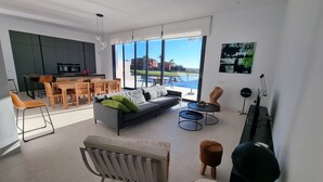 A modern living room with a sofa, coffee table, and chairs, a kitchen area with a dining table, and large windows offering a view of a body of water and buildings. - Moderne villa, met artistieke inrichting. dicht bij wandelpaden, zee, golfgebied (Vera)