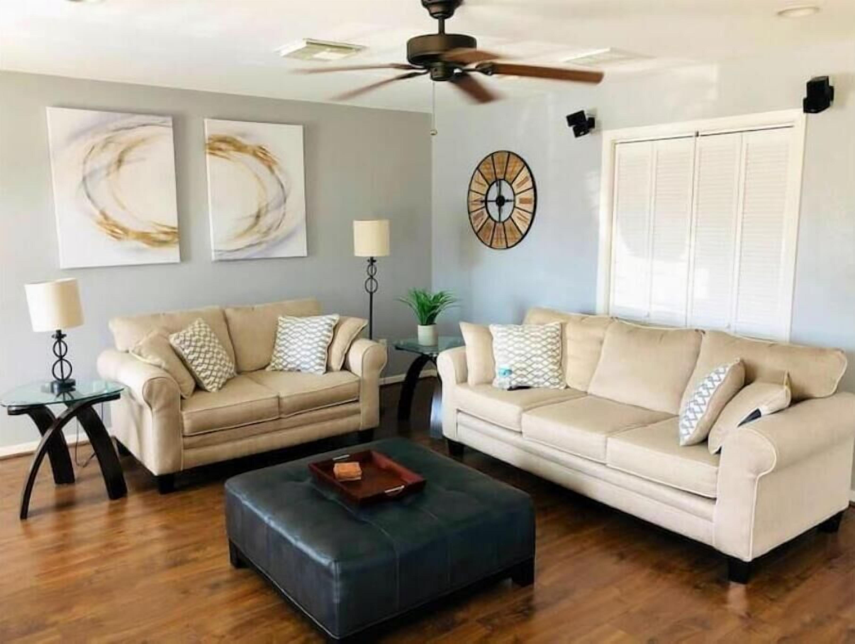 Una sala de estar con dos sofás color beige, un otoman oscuro, un ventilador de techo y un reloj de pared.
