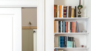 A bathroom with a white door, a mirror, and a sink with a faucet. Adjacent to the bathroom is a bookshelf with various books and a small lamp.