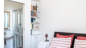 A hotel room with a bed, a bedside table, a mirror, a bathroom with a sink and shower, and a bookshelf.