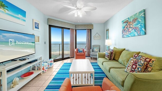 A living room with a TV, a ceiling fan, a sofa, a coffee table, and a view of the ocean through large windows.