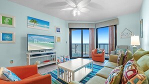 A living room with a TV, a ceiling fan, a sofa, a coffee table, and a view of the ocean through large windows.