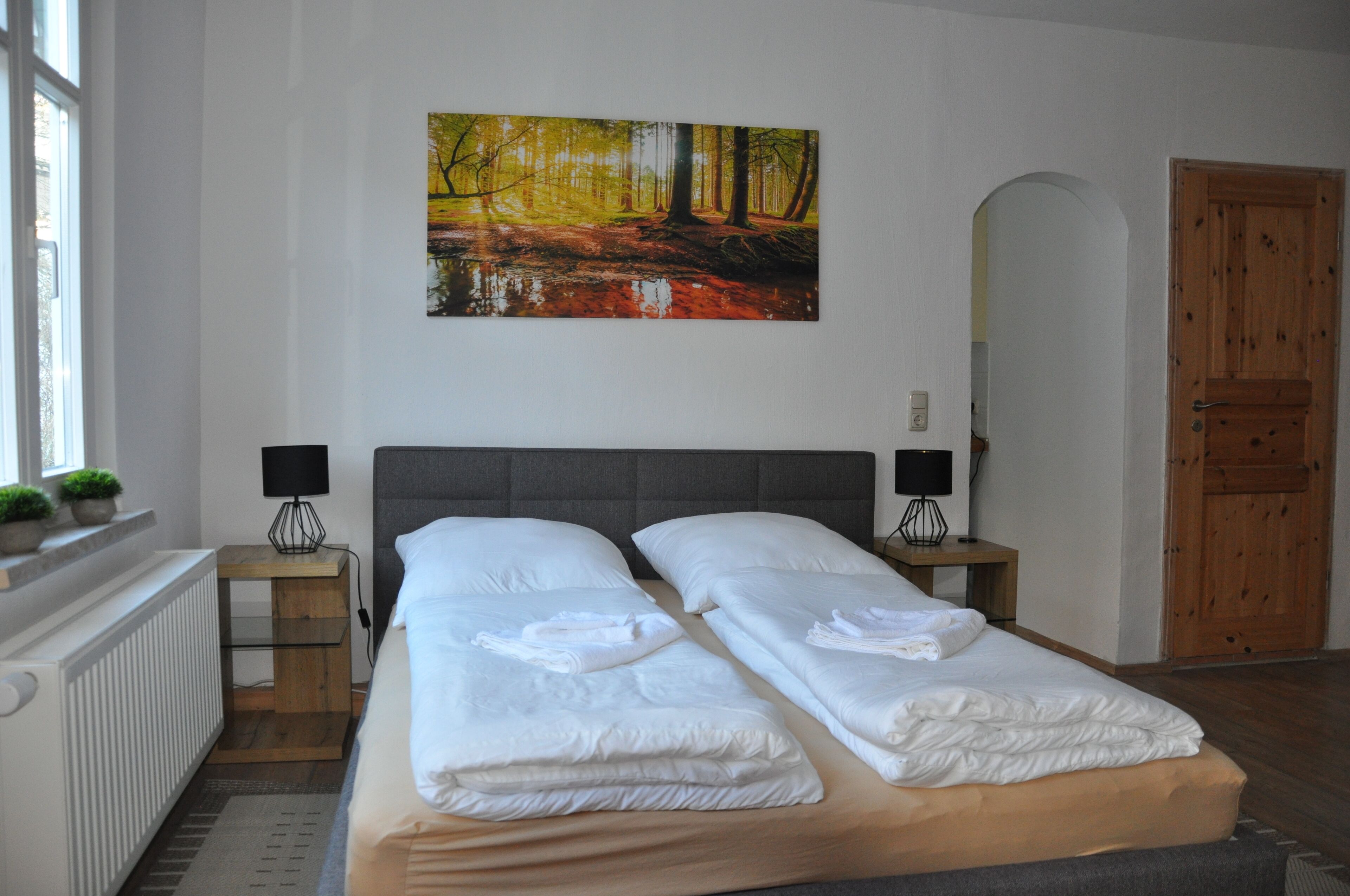 Une chambre d’hôtel avec un lit double, deux tables de chevet avec des lampes, un tableau représentant une scène de forêt, un radiateur et une porte en bois.