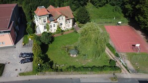 An aerial view of a large, historic building with a red-tiled roof, surrounded by lush greenery and a basketball court. - Ferienwohnung Gerberstein (Ruhla)