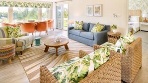 Ein geräumiges Wohnzimmer mit grauem Sofa, orangenen Sesseln und Korbmöbeln. Ein großes Fenster bietet Blick auf die grüne Umgebung.