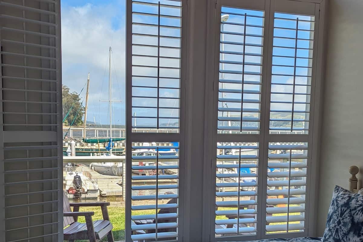 Une chambre avec une grande fenêtre donnant sur un port de plaisance, un lit recouvert d’une couvre-lit à motifs floraux et un fauteuil près de la fenêtre.