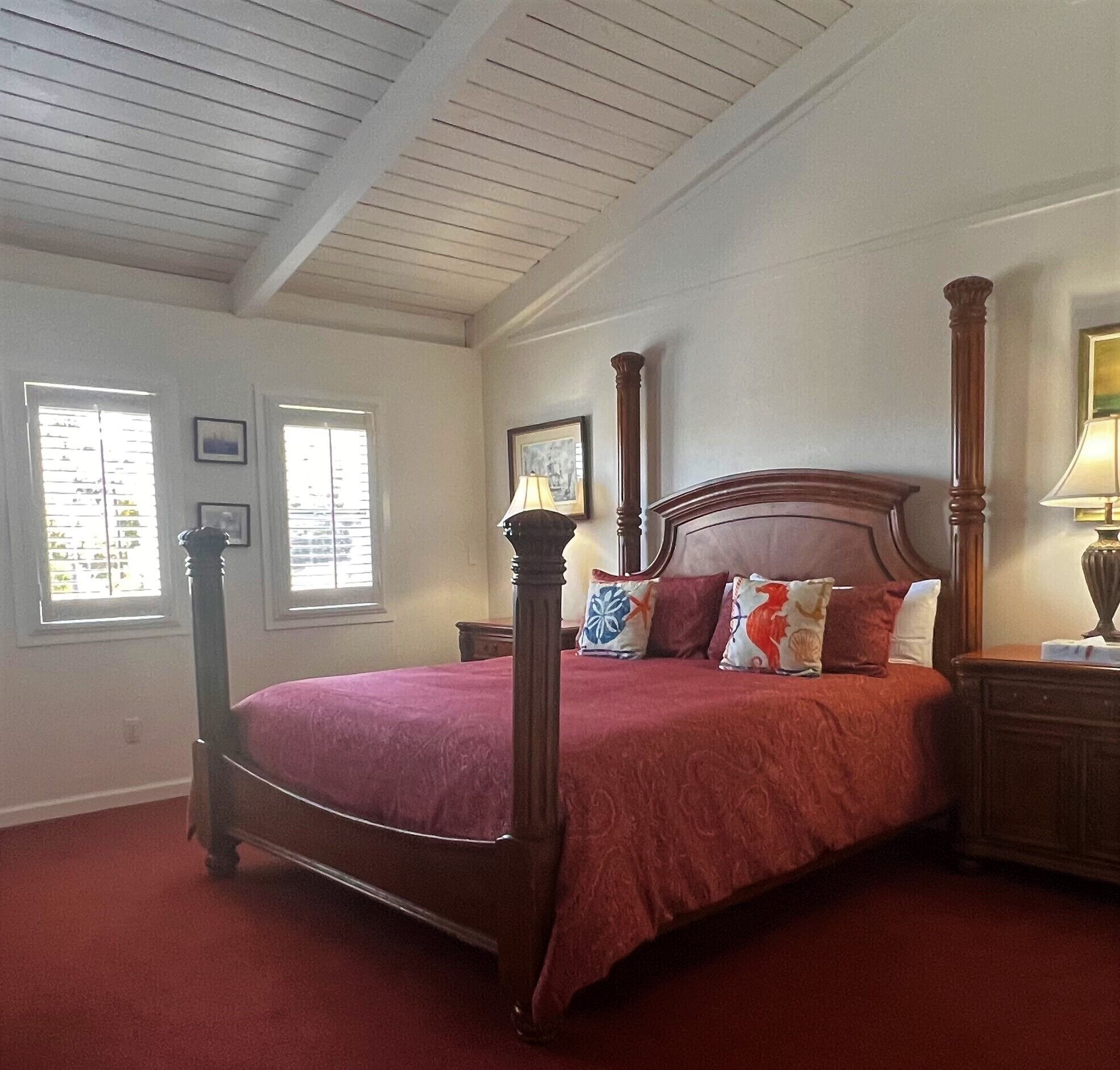 A four-poster bed with a red bedspread and decorative pillows, a nightstand with a lamp, and a window with blinds.