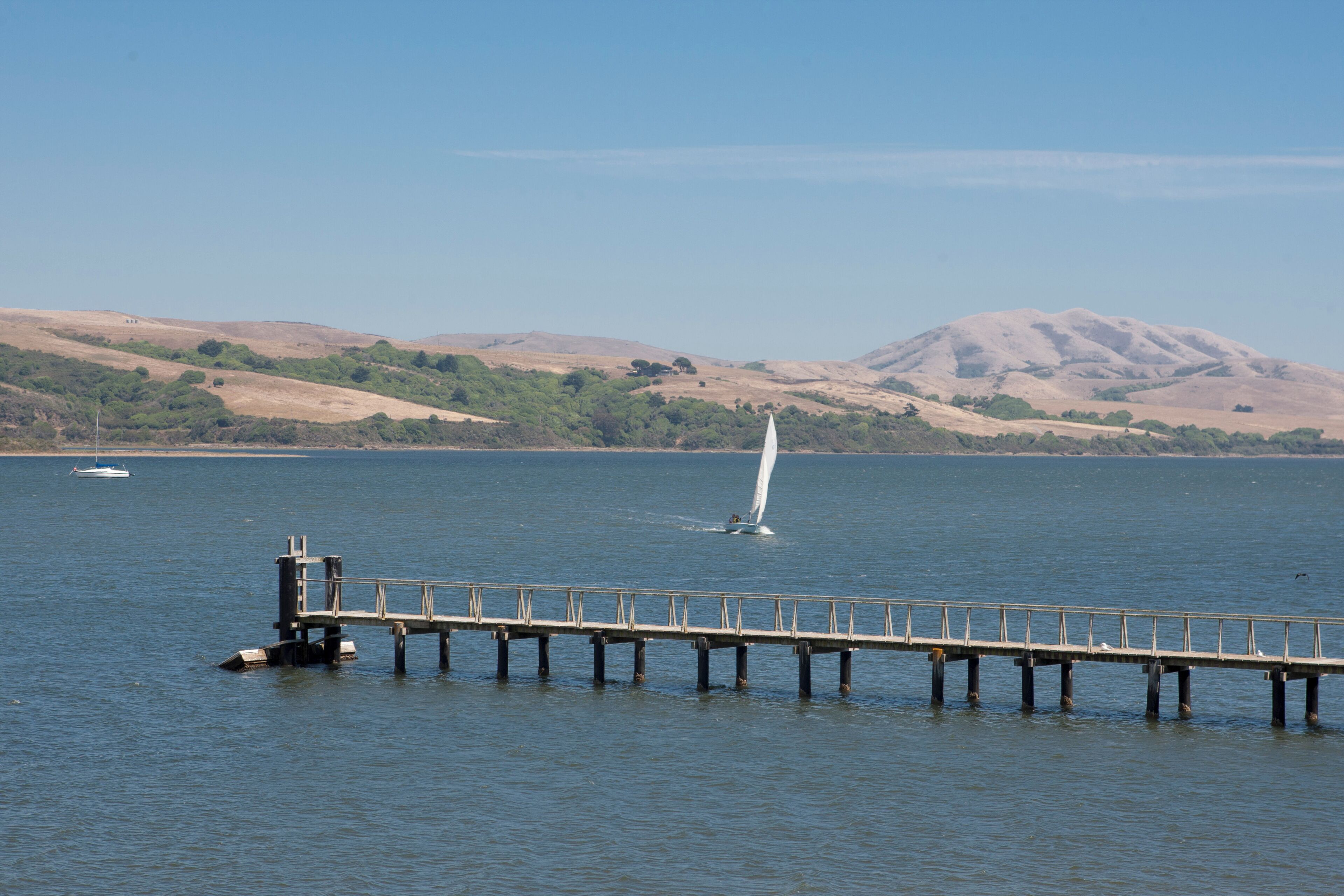 A pier extending into calm water with a sailboat in the distance and hills in the background.