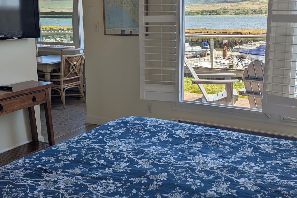 Une chambre avec un grand lit, un bureau et une chaise, donnant sur les bateaux et les collines.