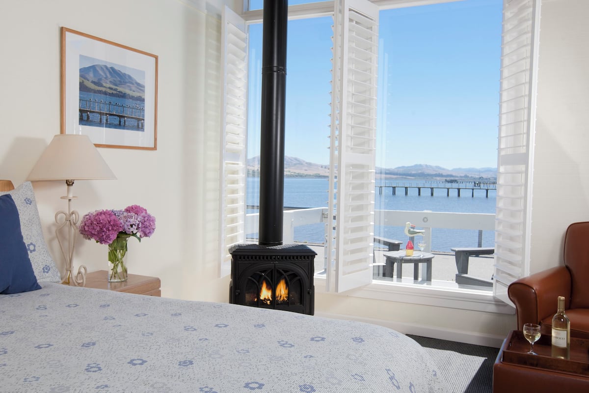 Une chambre à coucher avec un lit, une table de chevet avec une lampe et des fleurs, une cheminée et un balcon offrant une vue sur l’eau et les montagnes.