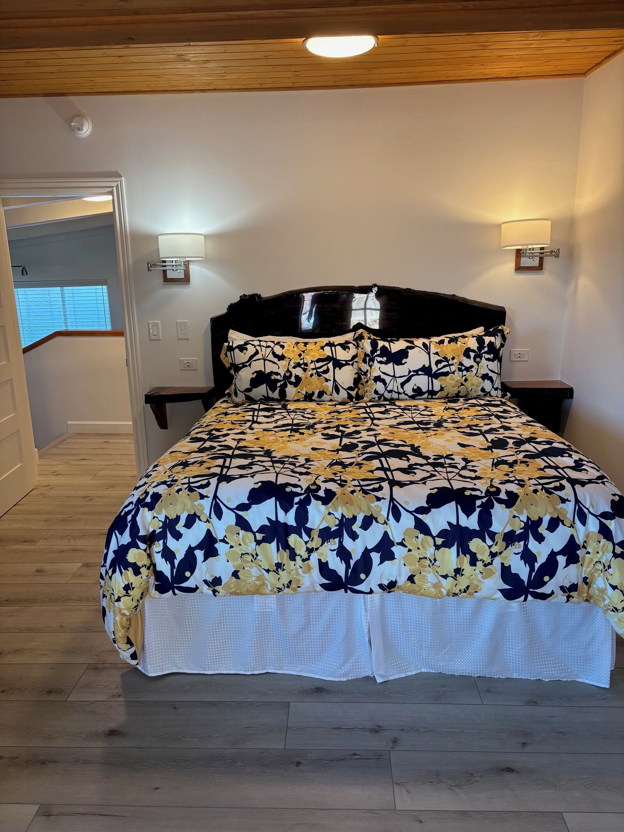 A bedroom with a bed, wooden floor, a ceiling light, a wall-mounted lamp, and a door leading to another room.