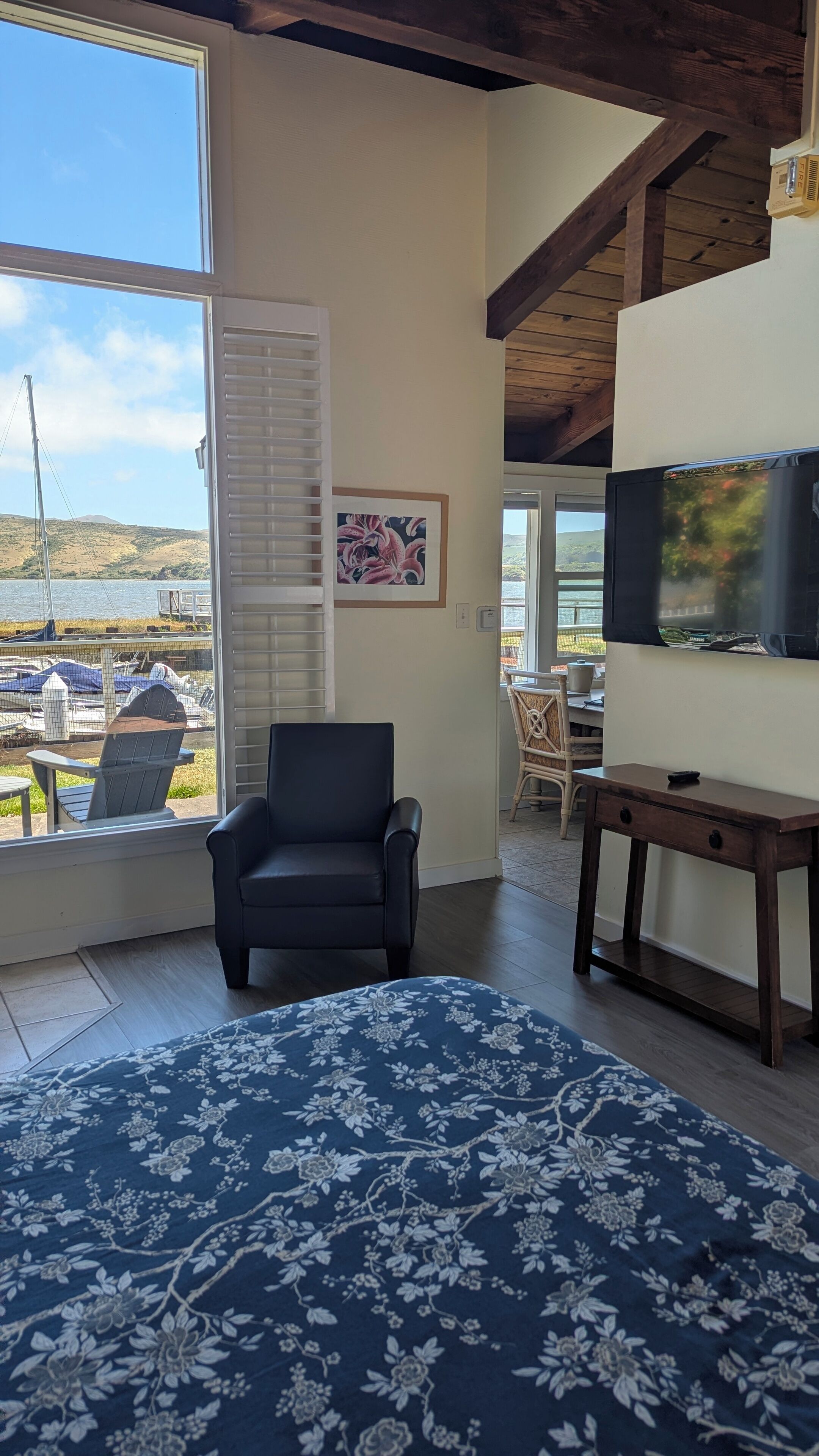A room with a large window, a bed with a floral bedspread, a chair, a TV on a wooden stand, and a picture on the wall.