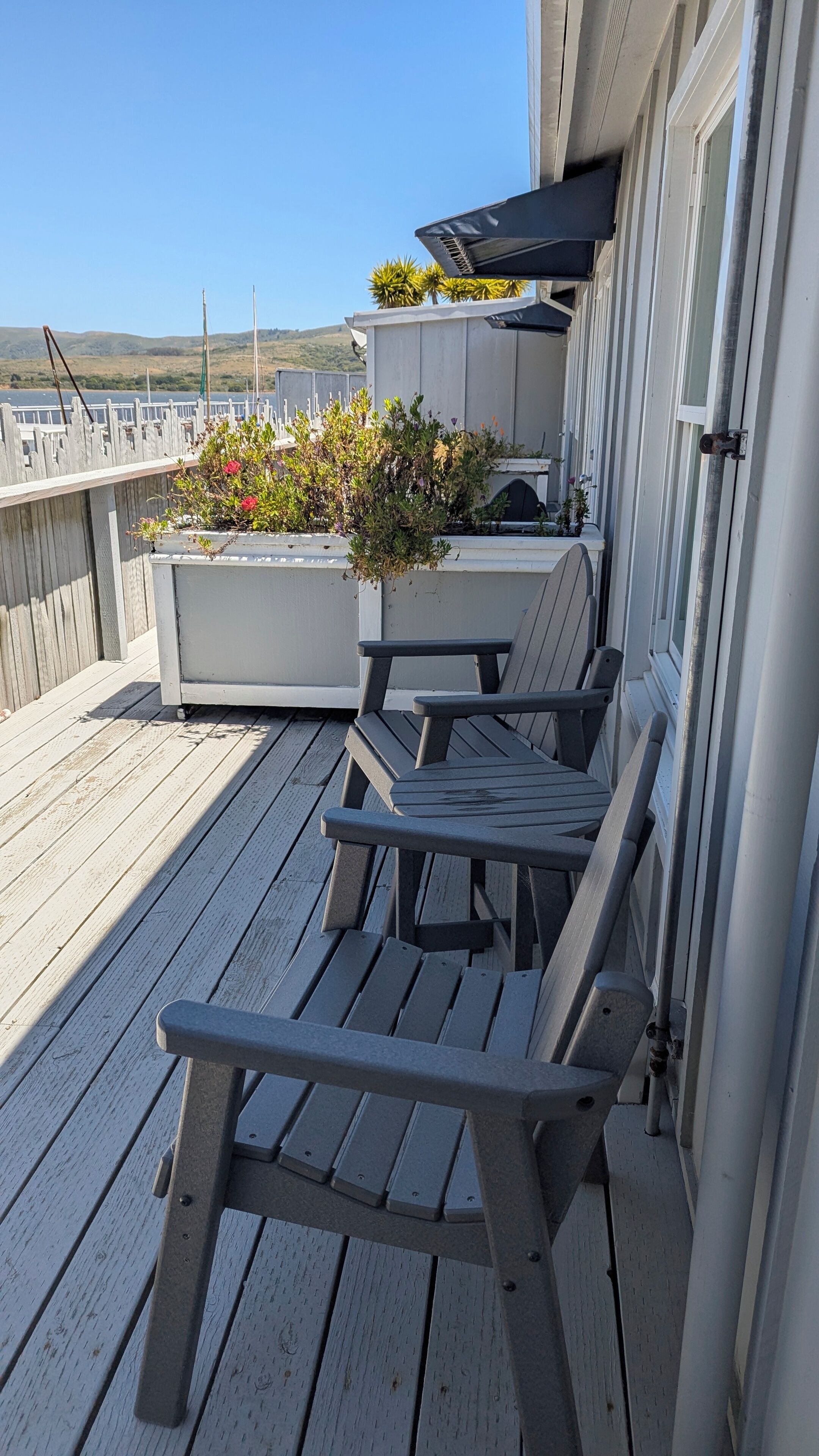 A wooden deck with two Adirondack chairs, a small planter with plants, and a view of a body of water and hills in the background.
