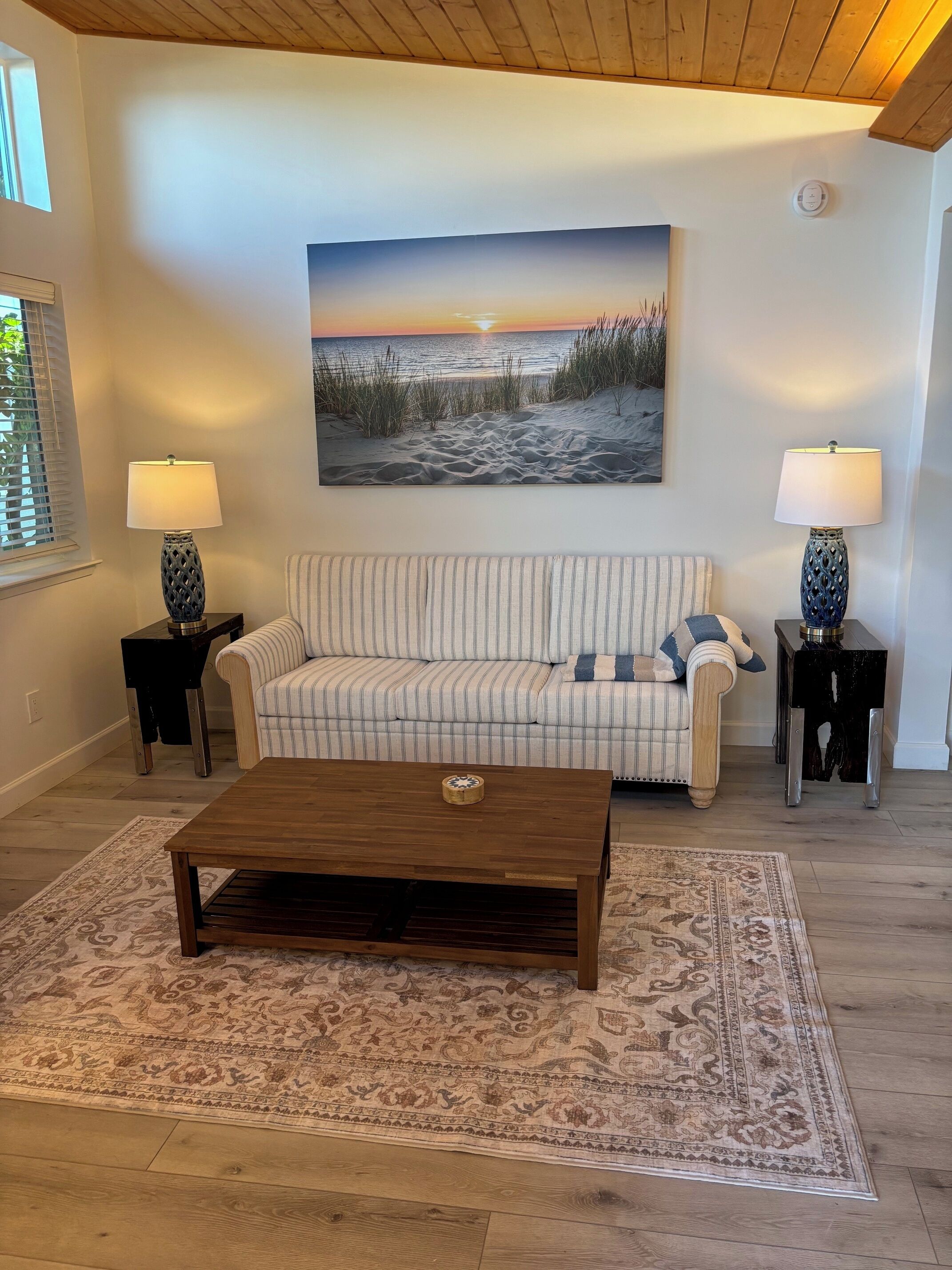 Uma sala de estar com mesa de centro de madeira, sofá listrado e duas luminárias em mesas laterais. Um quadro de praia está pendurado na parede acima do sofá.