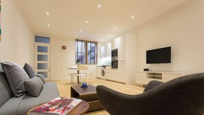 Ett modernt vardagsrum med en soffa, ett soffbord och en tidning. I bakgrunden syns ett kök med en tv och en tvättmaskin.