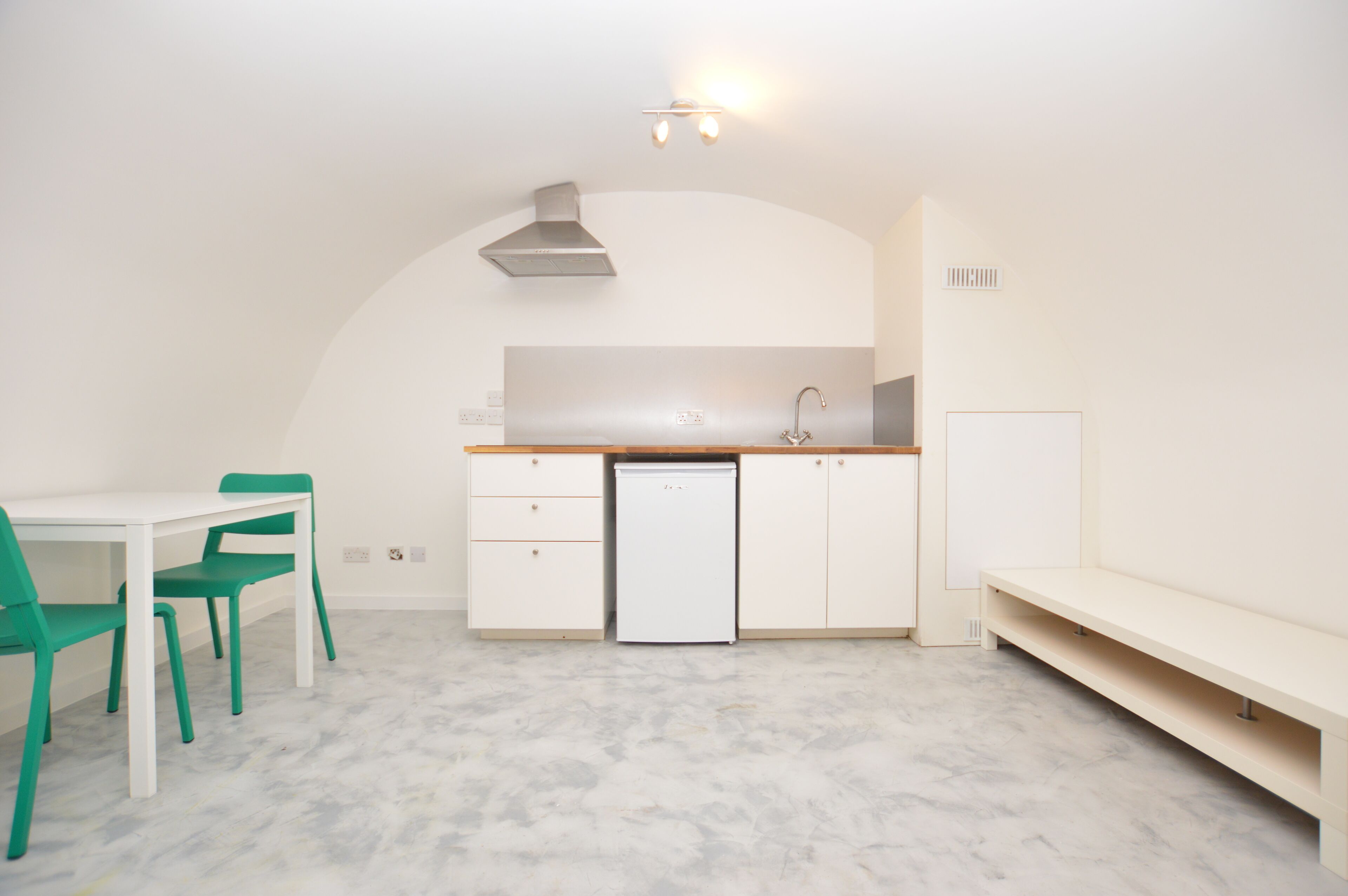 A compact kitchen with white cabinets, a white countertop, a sink, and a dishwasher. There is a dining table with green chairs and a bench.