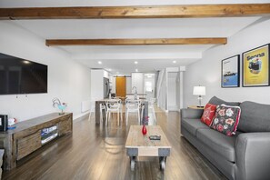 Un salon moderne avec un canapé gris, une table basse en bois et des chaises blanches. Un téléviseur est fixé au mur, et des tableaux encadrés ornent également le mur.