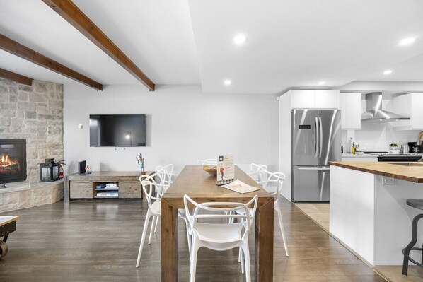 Une cuisine moderne dotée d’un réfrigérateur encastré, d’une table à manger en bois avec des chaises blanches et d’une cheminée en pierre.