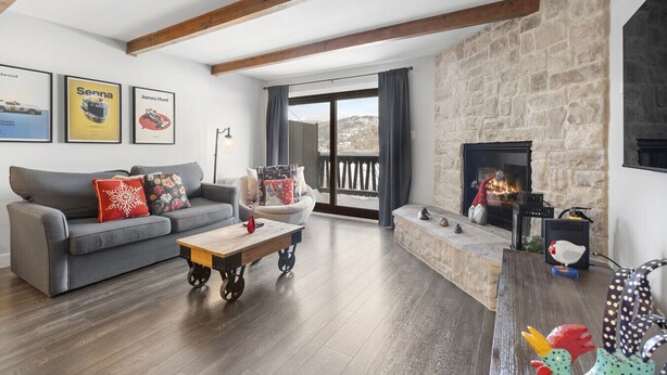 A living room with a stone fireplace, wooden beams, a grey sofa, and a view through a large window. - Luxury Condominium with Rustic Minimalist Style (Mont-Tremblant)