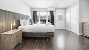 A modern bedroom with a wooden headboard, a large bed with white bedding, a nightstand with a lamp, and a view of the outdoors through a window with curtains. - Luxury Condominium with Rustic Minimalist Style (Mont-Tremblant)