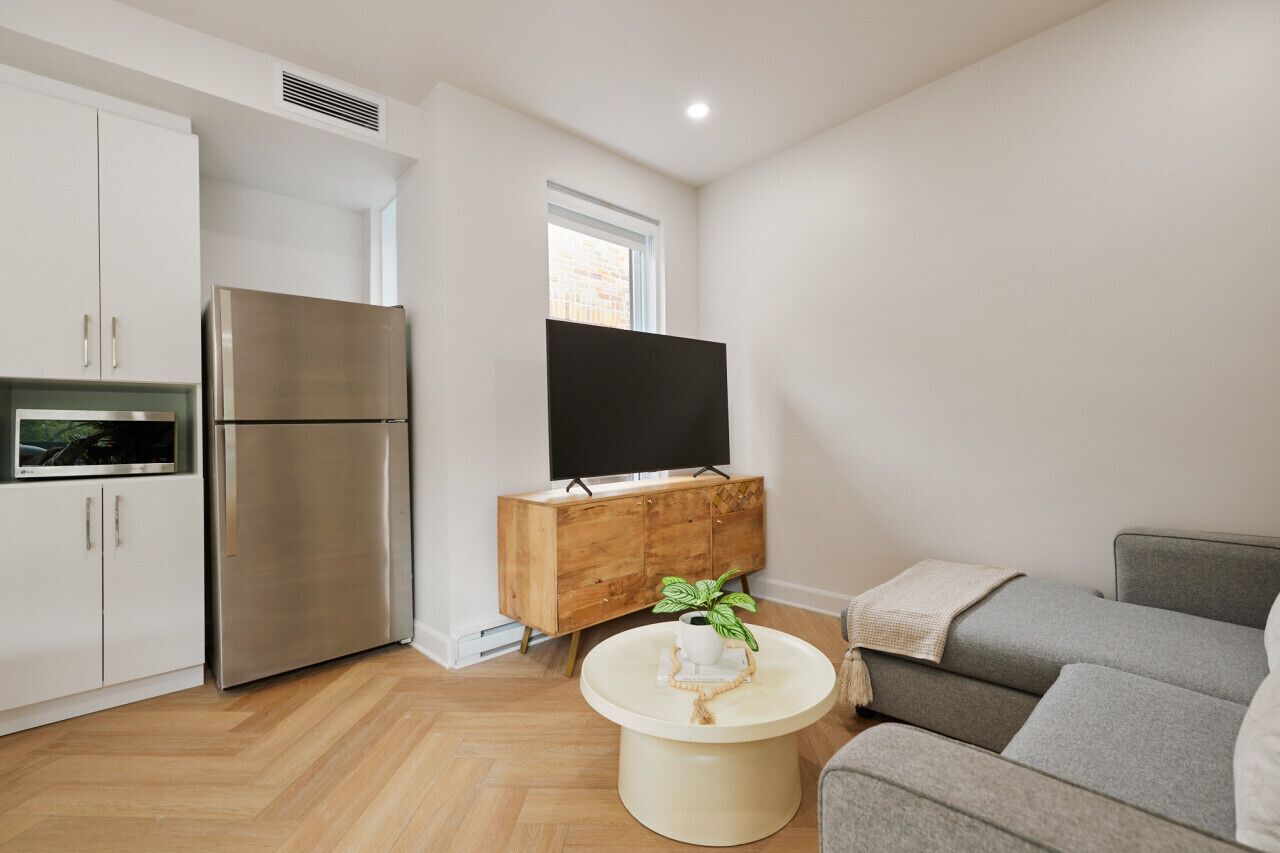 A modern kitchen with a stainless steel refrigerator, a wooden cabinet with a flat-screen TV, and a round beige table with a plant.