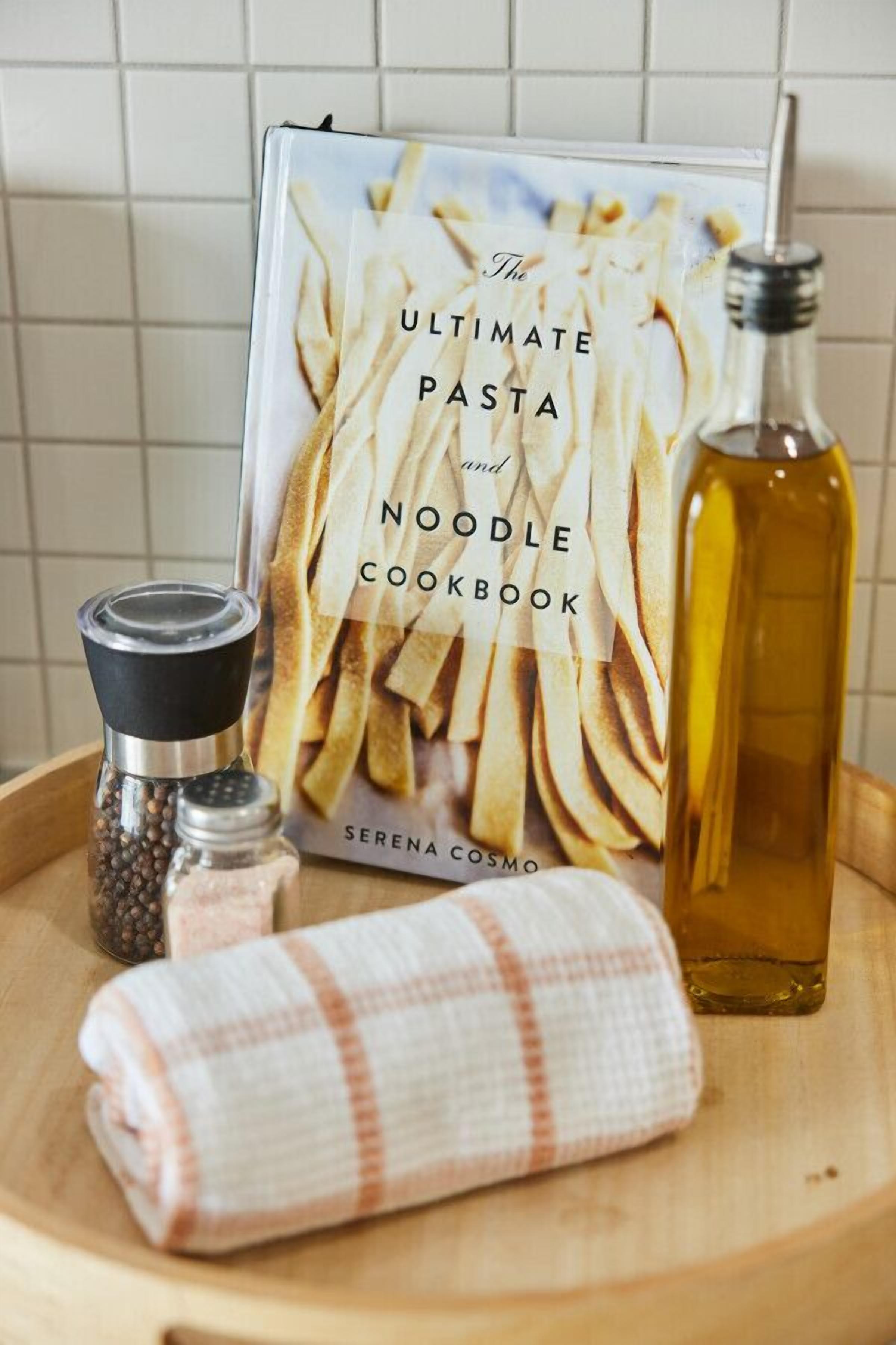 A cookbook titled "The Ultimate Pasta and Noodle Cookbook" by Serena Cosmos, a pepper grinder, a salt shaker, a bottle of olive oil, and a rolled-up cloth napkin on a wooden tray.
