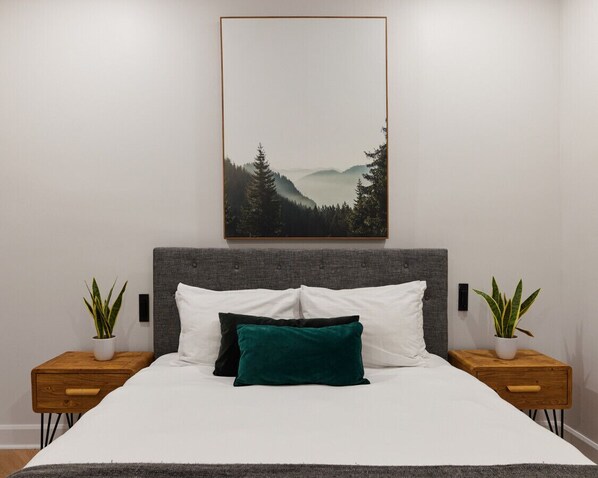 Un lit avec une tête de lit grise, des oreillers blancs et verts, et deux tables de chevet ornées de plantes en pot.