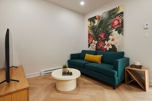 A living room with a teal sofa, a round coffee table, a TV on a stand, and a large floral painting on the wall.
