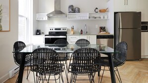 A modern kitchen with a glass dining table, black wicker chairs, white cabinets, stainless steel appliances, and a window providing natural light. - New and Cozy 3 bedroom in Le Plateau by Denstays (Montreal)