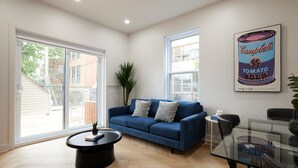 Ein modernes Wohnzimmer mit einem blauen Sofa, einem Glastisch und einem Esstisch mit Stühlen. Ein großes Fenster bietet einen Ausblick nach draußen, und an der Wand hängt ein Campbell's Tomato Soup-Poster.