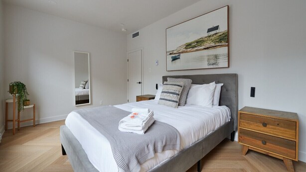Ein modernes Schlafzimmer mit einem großen Bett, einem hölzernen Nachttisch, einem kleinen Tisch mit Pflanze und einem Bild an der Wand.