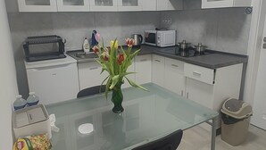 A compact kitchen with white cabinets, a glass dining table, and a black chair. There is a microwave, a kettle, and a trash bin visible. - Vu's Home - Apartment Junior - 40m2 (Hlavní město Praha)