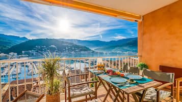 Een balkon met een tafel gedekt voor twee, met uitzicht op een jachthaven en stadsbeeld, met bergen op de achtergrond.