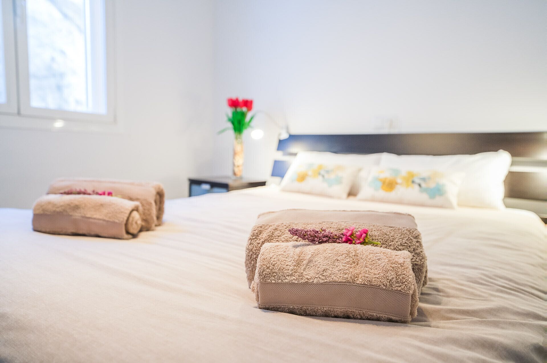 Een net opgemaakt bed met opgerolde handdoeken en een decoratieve bloemstelling op het nachtkastje.