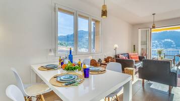 Een eetgedeelte met een witte tafel gedekt voor twee personen, uitzicht op zee en een balkon met bergzicht.