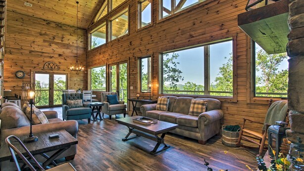 A spacious living room with wooden walls and large windows, furnished with sofas, a coffee table, and a fireplace. - Mtn Views, Fireplace, & 2-Story Wall of Windows!🔥 (Sevierville)