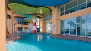 An indoor swimming pool with a colorful water slide, a lifeguard tower, and a view of the ocean through large windows.