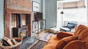 A cozy living room with a brick fireplace, a modern sofa, and a large window providing natural light. - 1 bedroom accommodation in Carlisle (Carlisle)