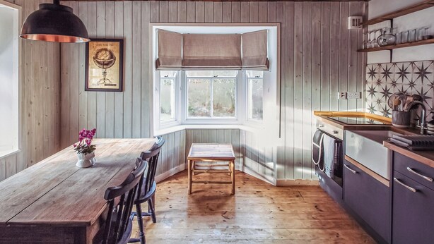 A kitchen with wooden walls, a dining area with a wooden table and chairs, a window with blinds, and a hanging light fixture. - 1 bedroom accommodation in Carlisle (Carlisle)